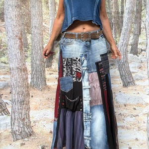 May include: A long patchwork skirt constructed from various denim panels, black fabric with white text, plaid fabric, and solid maroon sections. The skirt features a brown leather belt with a metal buckle and a frayed, multi-textured hemline.