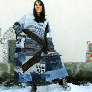 Op de afbeelding: Een lange, grijze en bruine hoodie met een patchwork-ontwerp met blauwe denim. De trui wordt gedragen door een persoon die in de sneeuw staat.