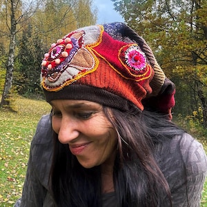 May include: A vibrant, handmade beanie hat with a patchwork design. The hat features a mix of textures and colours, including red, orange, brown, and beige. Embellishments include embroidered details and small, pink pom-poms. The hat is worn outdoors.