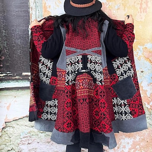 May include: A long coat with a striking pattern in red, black, and grey. The coat showcases detailed geometric designs and patchwork. It has a black collar and pockets, and is worn with a black hat.