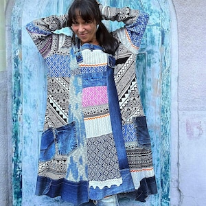 Op de afbeelding: Een tuniek met lange mouwen voorzien van een patchworkontwerp van blauwe spijkerstof en diverse zwart-wit-blauwe geometrische patronen. Het kledingstuk bevat meerdere spijkerstof zakken, een kraag met knoopsluiting en een uitlopende zoom.