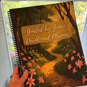Op de afbeelding: Een spiraalgebonden dagboekplanner met een omslag met een illustratie van een pad door een bos, met roze bloemen en de tekst "Guided by Grace Devotional Planner".