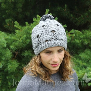 Puede incluir: Un gorro de punto gris con un pompón negro en la parte superior. El gorro tiene un patrón de encaje y lo lleva una persona con el pelo largo y castaño.