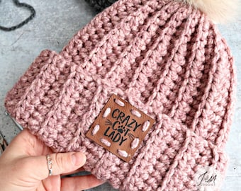 May include: A pink crocheted beanie with a faux fur pom pom and a brown leather patch that says "Crazy Cat Lady".