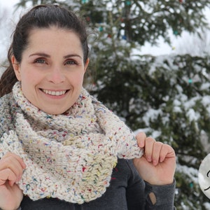 May include: A white, gray, and multi-colored crochet infinity scarf with a speckled pattern. The scarf is worn by a person in a snowy outdoor setting.