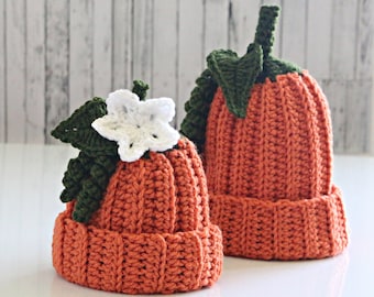 May include: Two crocheted pumpkin hats, one larger than the other. The smaller hat has a white crocheted flower and green leaves on top. Both hats are orange.