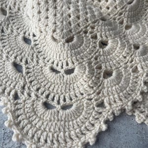 May include: A cream-colored crochet textile featuring a repeating fan-shaped pattern with intricate stitching. The design consists of layered, scalloped edges created with yarn, showcasing a detailed, openwork texture throughout the fabric.