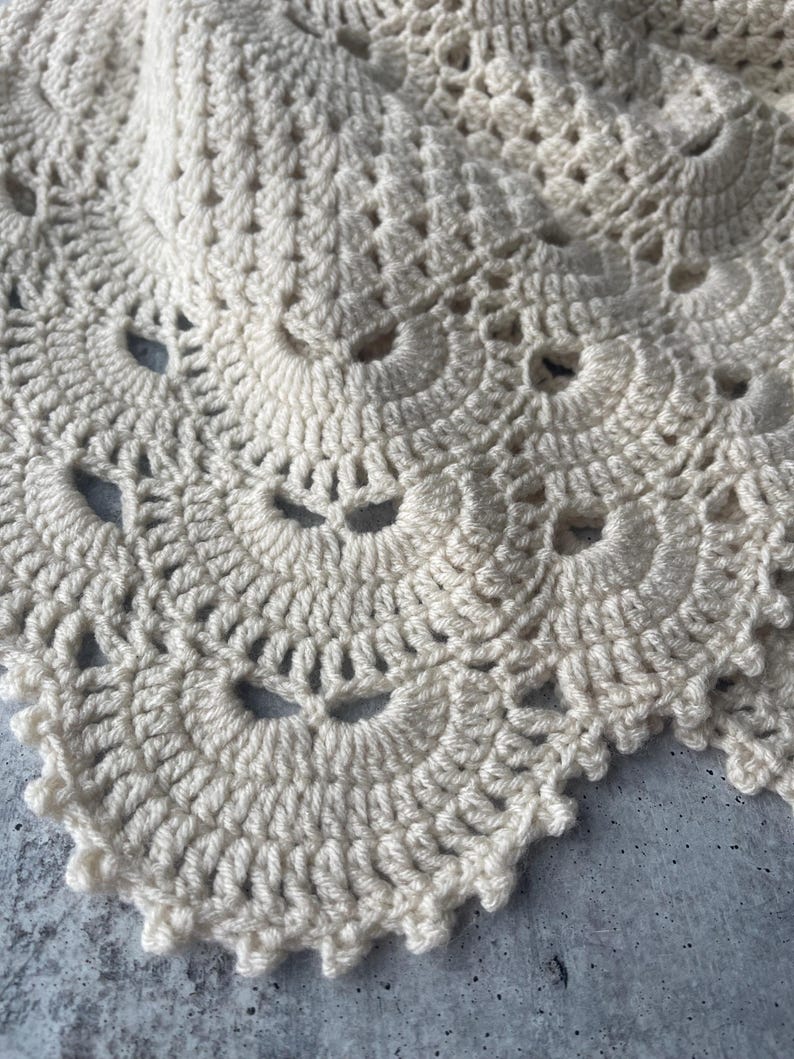 May include: A cream-colored crochet textile featuring a repeating fan-shaped pattern with intricate stitching. The design consists of layered, scalloped edges created with yarn, showcasing a detailed, openwork texture throughout the fabric.