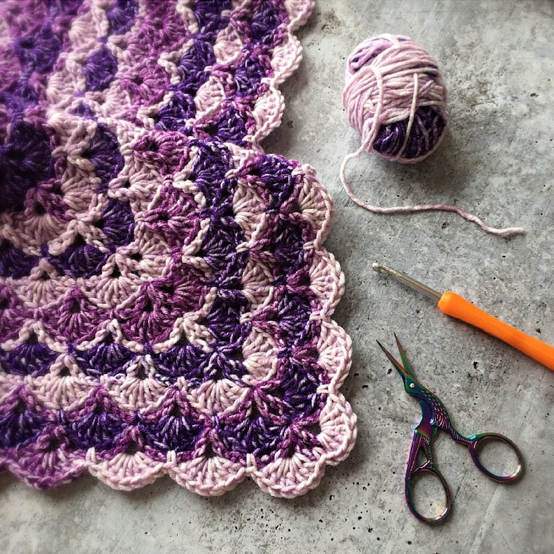 May include: A close-up of a crochet project in shades of purple and pink, with a scalloped edge. A ball of yarn, a crochet hook, and rainbow scissors are next to the project, suggesting a crafting theme.