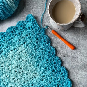 May include: A blue and green ombre crochet blanket with a crocheting hook and yarn ball in the background.