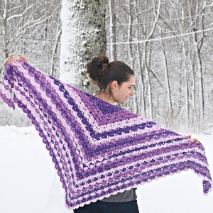 May include: A handmade, crocheted shawl in shades of purple and white. The shawl features a scalloped edge and a striped pattern. The woman is holding the shawl open, showcasing the design against a snowy backdrop.