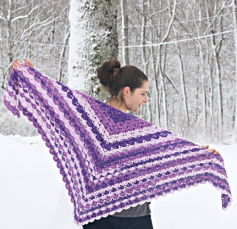 May include: A handmade, crocheted shawl in shades of purple and white. The shawl features a scalloped edge and a striped pattern. The woman is holding the shawl open, showcasing the design against a snowy backdrop.