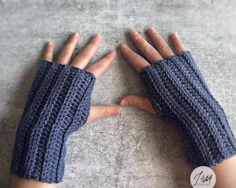Puede incluir: Un par de guantes sin dedos de color azul con un patrón de ganchillo de canalé vertical. Los guantes cubren la muñeca y la palma de la mano, dejando los dedos y el pulgar al descubierto. Se ve un pequeño logotipo circular con el texto JM CROCHET.