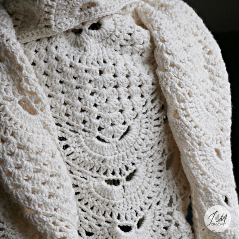 May include: An off-white crochet shawl featuring a repeating fan-like stitch pattern with openwork details. The fabric has a textured, knotted appearance throughout. A small circular logo in the bottom right corner reads JM CROCHET.