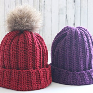 Peut inclure: Deux bonnets d'hiver en tricot, un rouge avec un pompon en fausse fourrure marron et un violet.