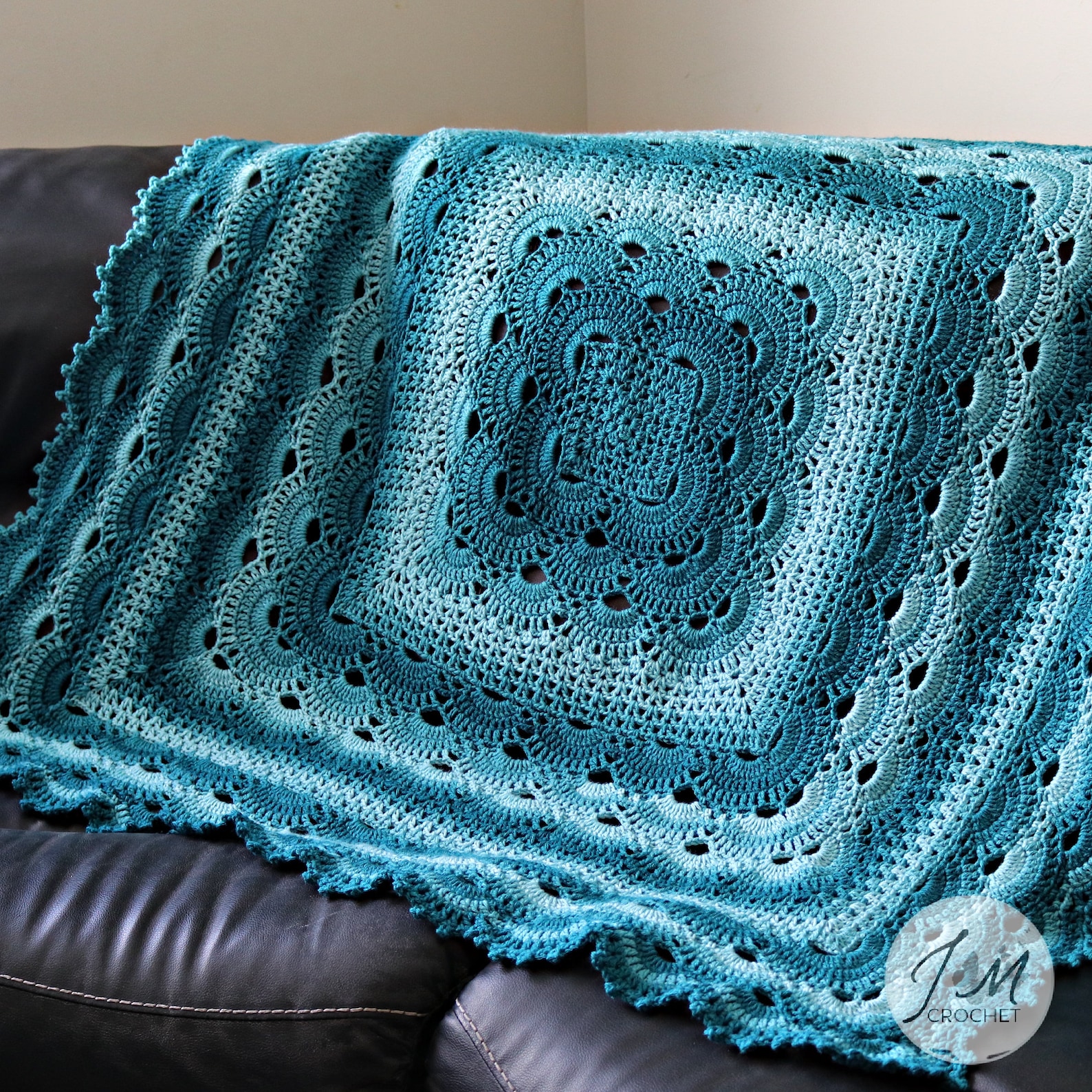 Crocheted blanket in rich teal ombré, displayed draped over a black leather couch with shell stitch detail and scalloped edging