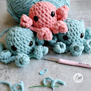 Op de afbeelding: Drie handgemaakte gehaakte octopussen in roze en blauwe tinten. De octopussen hebben zwarte knoopogen en gekrulde tentakels. Een haaknaald, steekmarkeerder en garen zijn ook zichtbaar.