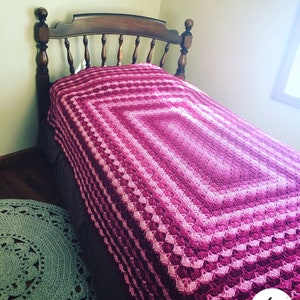 Puede incluir: Una manta de ganchillo rosa y morada con un borde festoneado está drapeada sobre un marco de cama de madera. La manta tiene un patrón repetitivo de cuadrados y está hecha con una variedad de tonos de rosa y morado.