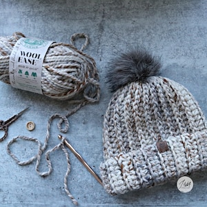 May include: A crocheted beanie hat in a beige and brown color with a faux fur pom pom on top. The hat is made with Lion Brand Wool Ease yarn, which is thick and quick to work with. The yarn ball and a crochet hook are also visible in the image.