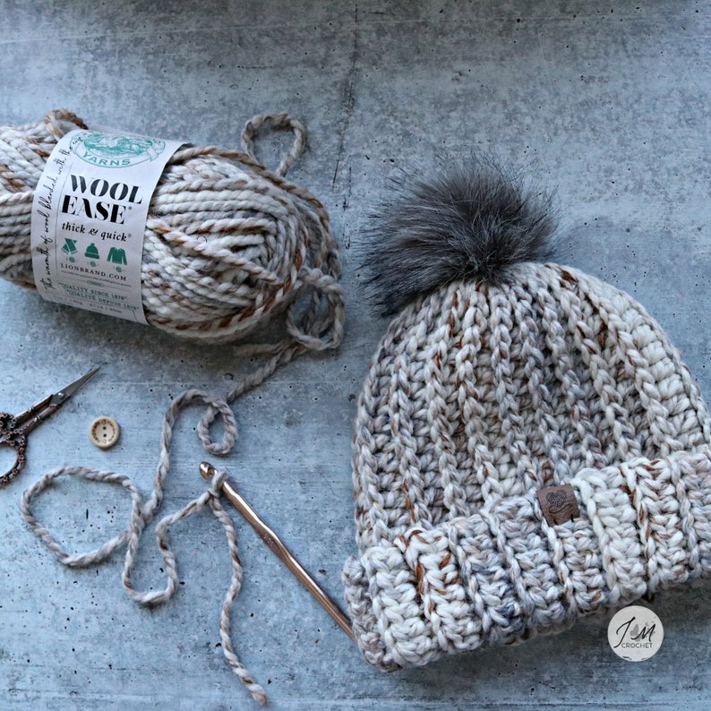 May include: A crocheted beanie hat in a beige and brown color with a faux fur pom pom on top. The hat is made with Lion Brand Wool Ease yarn, which is thick and quick to work with. The yarn ball and a crochet hook are also visible in the image.