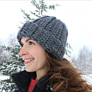 May include: A gray crocheted beanie hat with a ribbed texture. The hat is worn by a person with long brown hair, standing in a snowy outdoor setting.