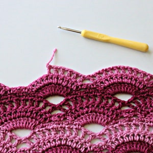 May include: A yellow crochet hook with a metallic tip positioned above a piece of fabric featuring a scalloped, wave-like pattern in varying shades of pink and magenta yarn, showing an unfinished edge with a loose pink thread.