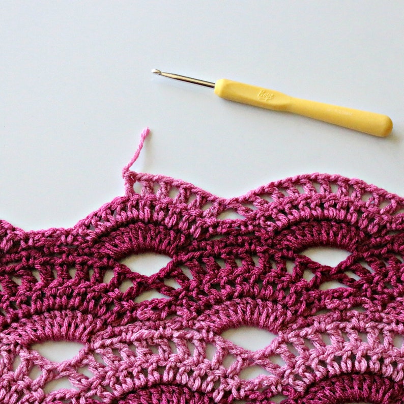 May include: A yellow crochet hook with a metallic tip positioned above a piece of fabric featuring a scalloped, wave-like pattern in varying shades of pink and magenta yarn, showing an unfinished edge with a loose pink thread.