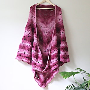 May include: A crochet shawl featuring an intricate fan stitch pattern in a gradient of pink and burgundy hues. The garment is displayed on a wooden hanger, showcasing a scalloped edge and an openwork design that transitions from light pink to deep red.