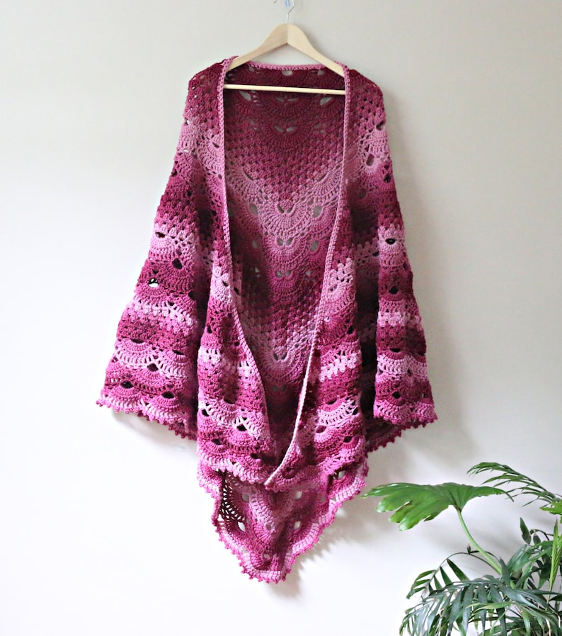 May include: A crochet shawl featuring an intricate fan stitch pattern in a gradient of pink and burgundy hues. The garment is displayed on a wooden hanger, showcasing a scalloped edge and an openwork design that transitions from light pink to deep red.