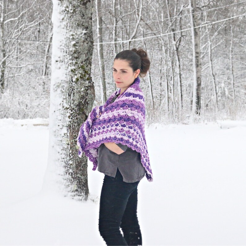 May include: A person is wearing a purple and white crocheted shawl in a snowy outdoor scene. The shawl has a scalloped edge and a striped pattern in shades of purple and white. The person is wearing a grey shirt and black trousers.