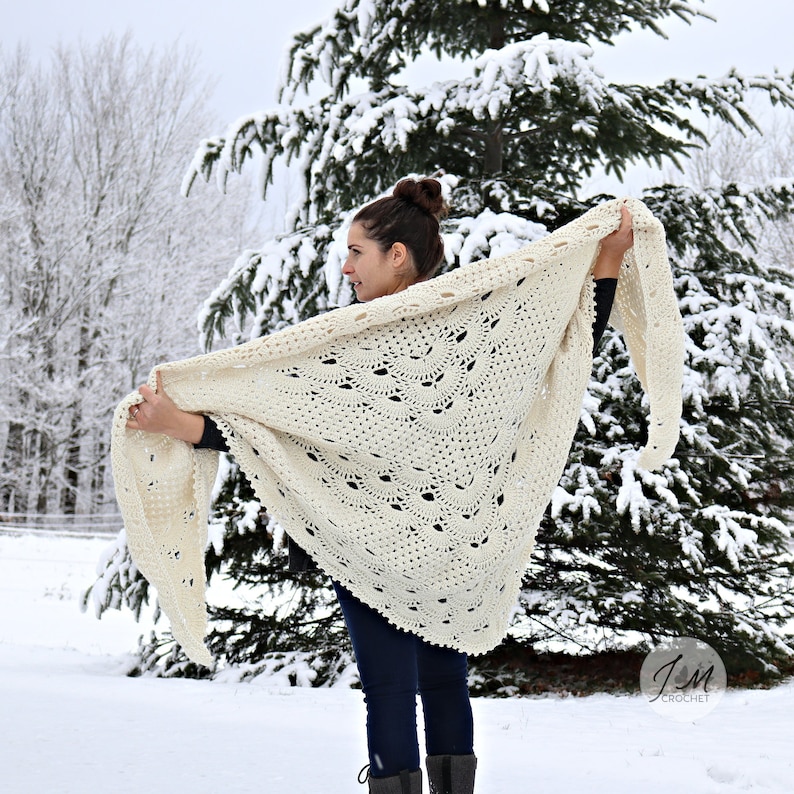 May include: A large, off-white triangular shawl featuring an intricate, repeating fan-shaped crochet pattern with a textured border. The accessory is held open to display the full scale of the detailed, hand-stitched yarn construction.