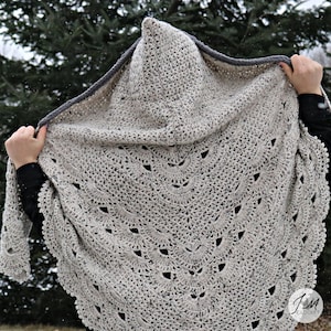 Può includere: Uno scialle all'uncinetto beige chiaro con cappuccio, con un bordo smerlato e ritagli decorativi. Il cappuccio è rifinito con un bordo grigio. Lo scialle è tenuto in alto dalle mani, mostrando l'intricato lavoro a punto e il design.