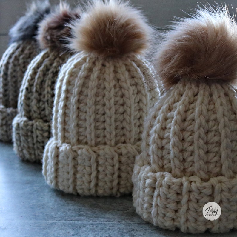 May include: Four crocheted hats with faux fur pom poms. The hats are in various shades of beige and brown. The hats are lined up in a row on a gray surface.