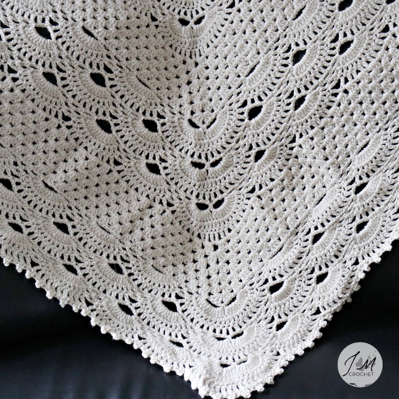 May include: An off-white crochet blanket featuring a repeating fan-shaped stitch pattern with openwork details. The bottom right corner displays a circular logo with the text JM CROCHET and a small butterfly icon.