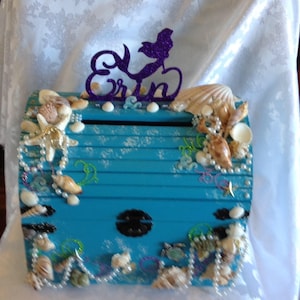 May include: A teal blue wooden treasure chest decorated with seashells, pearls, and a mermaid silhouette. The chest has a slot for inserting cards or money and is perfect for a mermaid-themed party or event.
