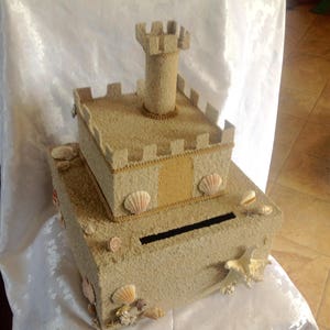 May include: A sand-textured box shaped like a castle, adorned with seashells and a central tower. The box has a slot for inserting items. The design incorporates a light beige color scheme with gold trim.