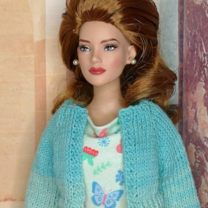 May include: A doll with auburn hair styled in a bouffant, wearing small pearl stud earrings, a light blue knitted bolero cardigan, and a light green sleeveless dress featuring a colourful pattern of butterflies, flowers, and small green leaves.