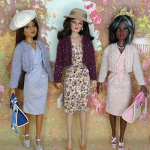 May include: Three dolls standing side by side. The left doll wears a white beret, blue cardigan, and blue dress with a blue handbag. The centre doll wears a straw hat, purple cardigan, and floral dress. The right doll wears a pink cardigan and dress.