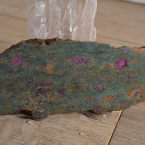 May include: A green and brown rock with pink ruby inclusions. The rock is sitting on a wooden surface.