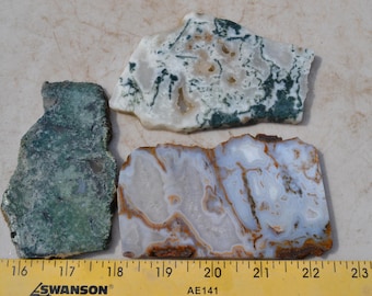 May include: Three irregularly shaped, polished stone slabs. One is green, one is white with green veining, and the third is white and blue with brown banding. A yellow ruler with black markings is at the bottom.