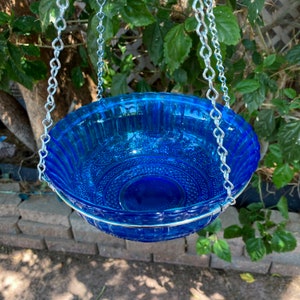 May include: A blue glass bird bath hanging from a silver chain. The bath has a decorative pattern and is suspended from a metal ring.