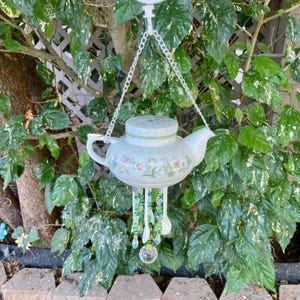 May include: A white ceramic teapot wind chime with a floral design, hanging from a white chain. The teapot has a lid and is adorned with colorful flowers. Green and clear beads and spoons dangle below, with a crystal ball at the bottom.