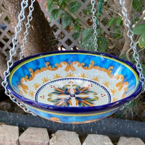 May include: A hanging ceramic bird bath with a vibrant blue and white floral design. The bowl is suspended by silver chains and features ornate gold accents. The bird bath is a decorative outdoor item.
