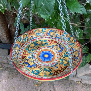 May include: A hanging ceramic bowl with a vibrant floral design in blue, yellow, and red. The bowl is suspended by silver chains, showcasing a colorful, decorative piece for outdoor use. The bowl's rim is red, and the interior features a central blue flower.