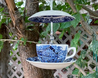 May include: A unique bird feeder crafted from a teacup, saucer, and a decorative plate, all featuring a blue and white pastoral design. The feeder is suspended from a tree branch by a silver metal rod. The teacup is approximately 4 inches in diameter.