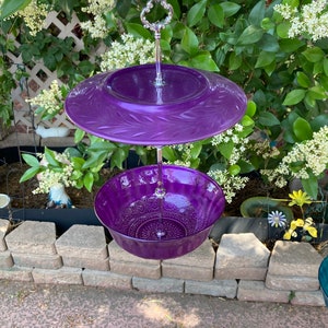 May include: A two-tiered purple serving dish with a decorative floral design. The top tier is a flat plate, and the bottom tier is a bowl. The dish is suspended by a silver metal handle, perfect for displaying desserts or appetizers.