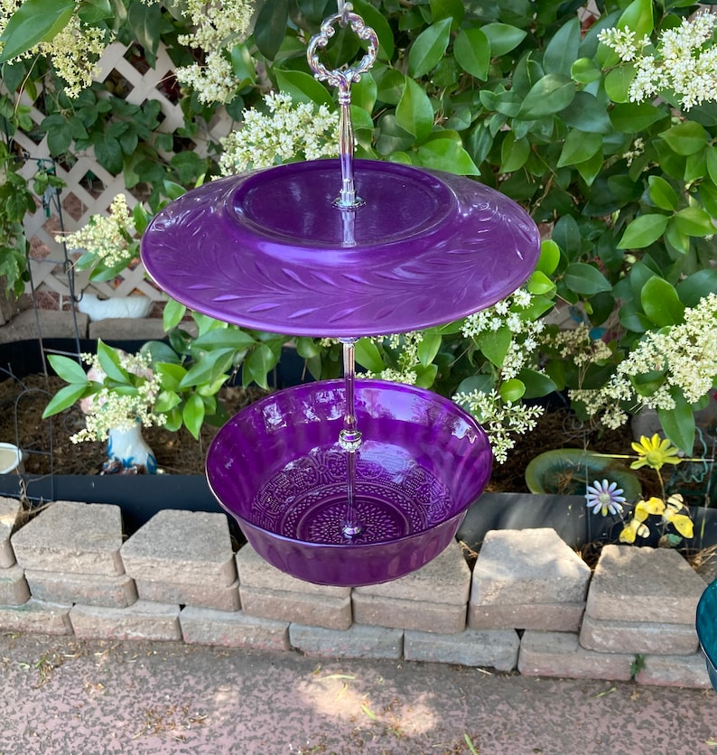 May include: A two-tiered purple serving dish with a decorative floral design. The top tier is a flat plate, and the bottom tier is a bowl. The dish is suspended by a silver metal handle, perfect for displaying desserts or appetizers.