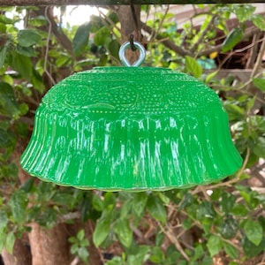 May include: A vibrant green glass lampshade with a decorative pattern, suspended from a metal ring. The lampshade has a flared, scalloped edge and is surrounded by green foliage. The glass has a glossy finish.