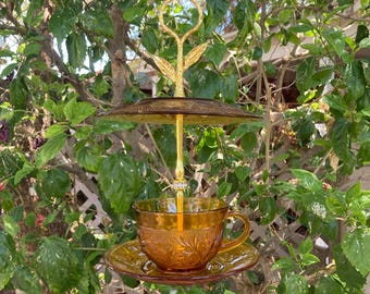 May include: A decorative, amber-colored glass bird feeder with a teacup and saucer design. The feeder has a gold-colored metal frame with a decorative top and a detailed, embossed pattern on the cup and saucer. The feeder is suspended among green leaves.