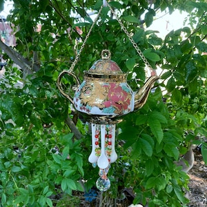 May include: A decorative hanging teapot wind chime featuring a white ceramic body with gold trim and floral patterns in pink, green, and gold. The teapot is suspended by a gold chain and features dangling white spoons and colorful glass beads.